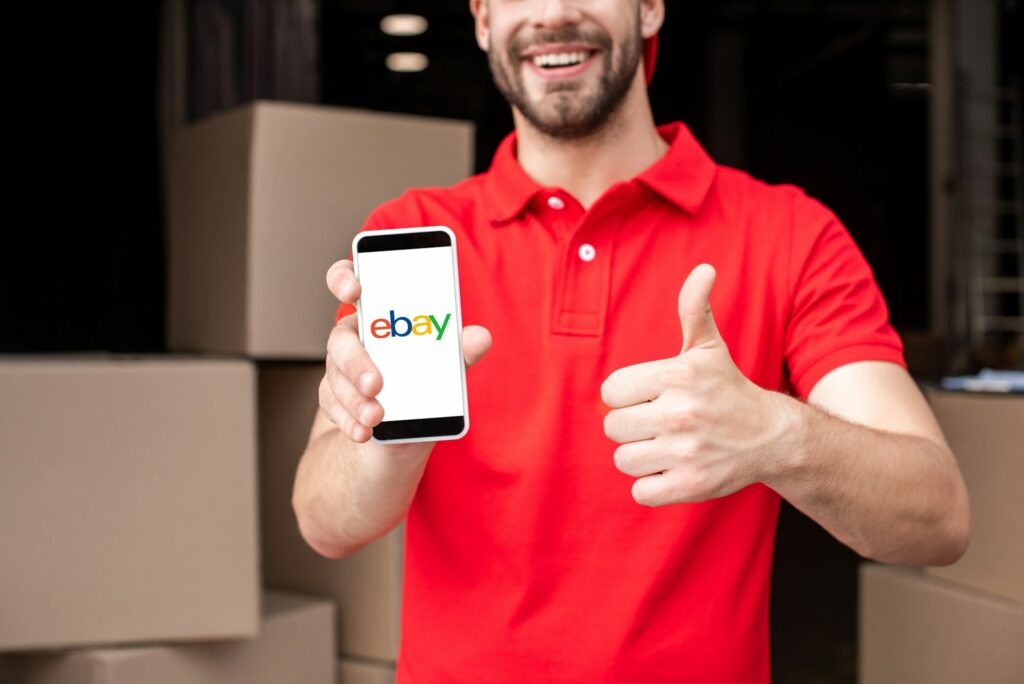 partial view of cheerful delivery man with smartphone with ebay logo on screen showing thumb up